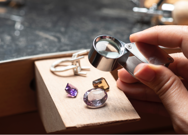 A hand holding a magnifying glass looking at jewels.