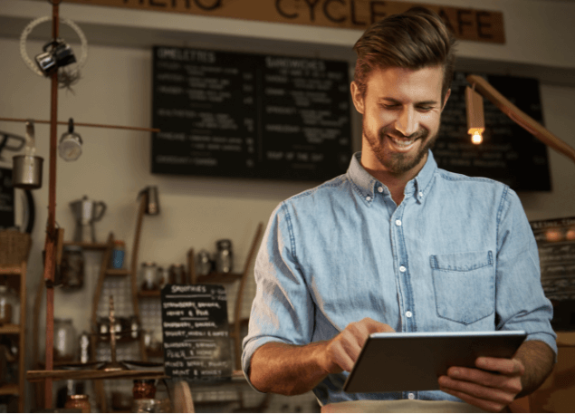 Man standing in coffee shop smiling and looking at a tablet.